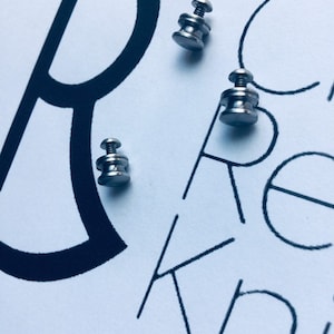 May include: Three silver metal knobs with a screw on top. The knobs are on a white surface with black lettering that says "Create Knit".