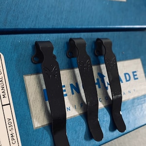 May include: Three black metal pocket clips for a knife. The clips are attached to a blue box with the text "Benchmade Knife Company" printed on it.
