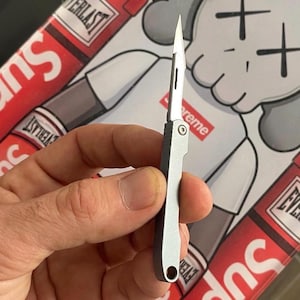 May include: A small, silver folding knife with a sharp blade. The knife is open, revealing the blade. The handle has a simple, rectangular design with a hole at the base. The background features a colorful design.