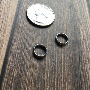 May include: Two small, silver-colored metal rings and a quarter coin are displayed on a dark brown wooden surface. The rings appear to be made of a shiny metal. The quarter coin is silver and shows a profile of a person.
