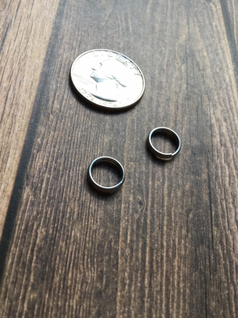 May include: Two small, silver-colored metal rings and a quarter coin are displayed on a dark brown wooden surface. The rings appear to be made of a shiny metal. The quarter coin is silver and shows a profile of a person.
