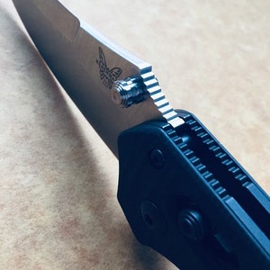 Könnte beinhalten: Nahaufnahme eines schwarzen Klappmessers mit einer teilweise geöffneten Klinge. Die Klinge hat eine silberne Oberfläche und ein Schmetterlingslogo. Der Griff hat eine strukturierte Oberfläche und eine gezackte Kante.