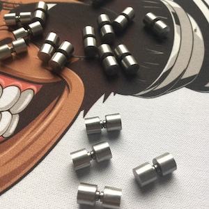 Könnte beinhalten: Nahaufnahme einer Gruppe silberner Metall-Druckknöpfe. Die Druckknöpfe sind in einem zufälligen Muster auf einer weißen Oberfläche mit einem Cartoon-Design angeordnet.