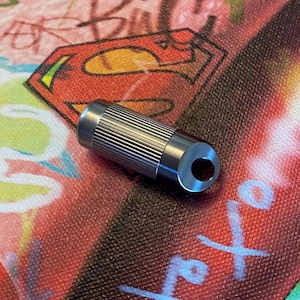 May include: A cylindrical, silver-colored metal object with a ridged design. The object has a hole at one end and is set against a colorful background featuring a Superman logo.