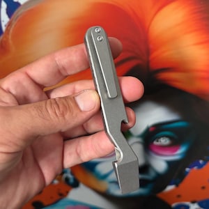 Könnte beinhalten: Ein mattgraues Metall-Multitool mit flachem, länglichem Körper und einem Taschenclip, der mit zwei Schrauben befestigt ist. Das Werkzeug verfügt über eine Flaschenöffner-Aussparung an der Seite und eine flache, abgewinkelte Hebelspitze.