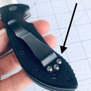 May include: A black pocket knife with a black clip and a textured handle. The clip is attached to the handle with four silver screws.