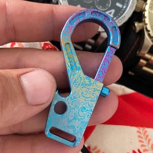 以下が含まれることがあります： 装飾的な模様のマルチカラーカラビナ。カラビナは主に青色で、紫と黄色のアクセントがあります。ボトルオープナーと小さなループが付いています。カラビナは人の手で持たれています。