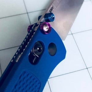Puede incluir: Un cuchillo de bolsillo azul con una hoja plateada y tornillos morados y azules. La hoja está parcialmente abierta y el cuchillo está sobre una superficie de azulejos blancos.