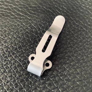 K&ouml;nnte beinhalten: Ein silberner Metall-Taschenclip mit zwei L&ouml;chern an der Seite und einem Schlitz in der Mitte.