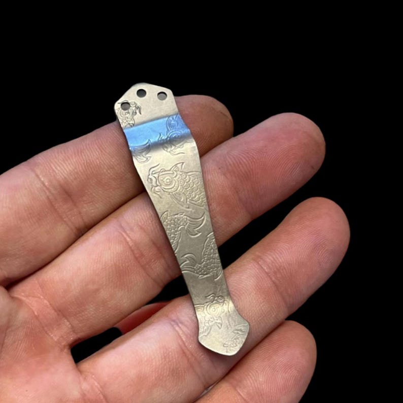 May include: A silver pocket clip with an engraved design of koi fish. The clip has three holes at the top for attaching to a pocket.
