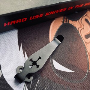 May include: Black metal pocket clip with a skull design. The clip is designed to attach to a pocket knife or other tool. The clip is made of durable metal and has a smooth finish.