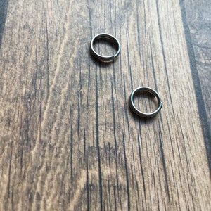May include: Two small, silver-colored split key rings are displayed against a textured, brown wooden surface. The rings are circular with a split for adding keys or other small items. The background is a natural wood grain.