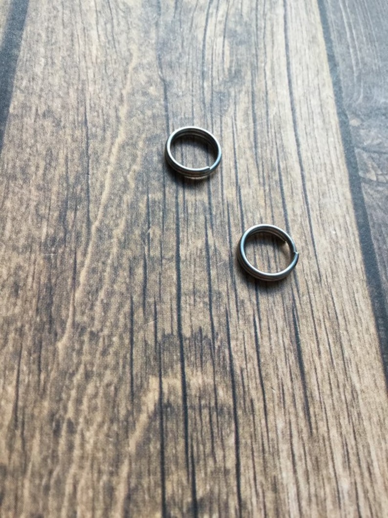 May include: Two small, silver-colored split key rings are displayed against a textured, brown wooden surface. The rings are circular with a split for adding keys or other small items. The background is a natural wood grain.