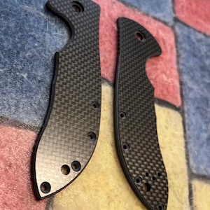 May include: Two carbon fiber knife scales with a woven pattern. The scales are dark gray and have several holes for screws. The scales are curved and have a smooth, polished finish. The knife scales are designed for a folding knife.