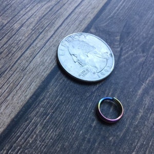 May include: A small, iridescent rainbow-colored ring next to a U.S. quarter coin. The ring appears to be made of metal and has a smooth, polished finish. The quarter is silver and shows the profile of a person.