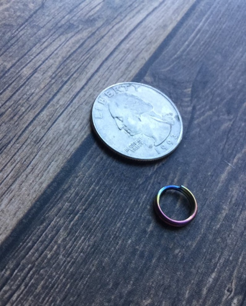 May include: A small, iridescent rainbow-colored ring next to a U.S. quarter coin. The ring appears to be made of metal and has a smooth, polished finish. The quarter is silver and shows the profile of a person.