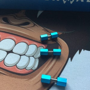 Könnte beinhalten: Drei kleine, hellblaue, hantelförmige Objekte mit silberfarbenen Verbindungsstangen. Die Objekte sind auf einer gemusterten Oberfläche angeordnet. Das Wort "BENCHMADE" ist im Hintergrund sichtbar.