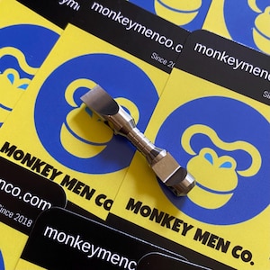 May include: A silver metal tool with a flat head and textured handle rests on business cards. The cards have a yellow background with a blue monkey logo and the text "MONKEY MEN Co" and "monkeymenco.com".