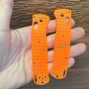 Puede incluir: Dos cachas de cuchillo de color naranja brillante con un patrón texturizado en forma de rejilla. Cada cacha tiene varios agujeros para tornillos y una forma curva. Las cachas se sostienen en una mano sobre un fondo de madera.