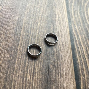May include: Two silver-colored split key rings are displayed on a wooden surface. The rings are circular with a split for attaching keys or other items. The wood grain is visible.