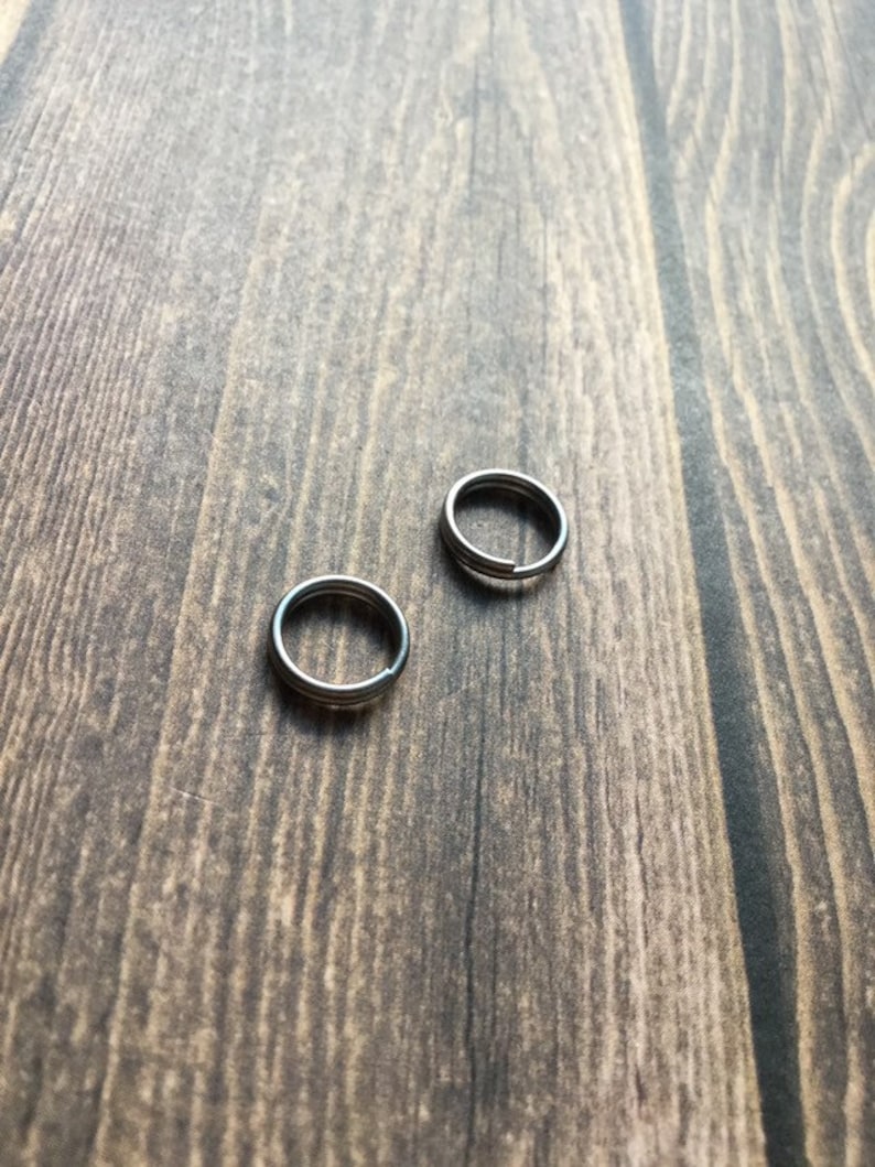 May include: Two silver-colored split key rings are displayed on a wooden surface. The rings are circular with a split for attaching keys or other items. The wood grain is visible.