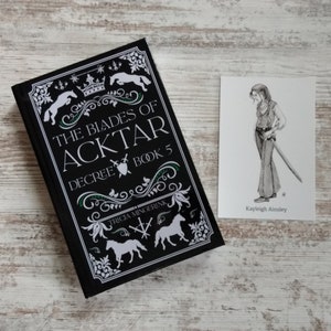 K&ouml;nnte beinhalten: Schwarzes Buch mit wei&szlig;em Text und Illustrationen. Der Titel lautet "The Blades of Aktar, Decree Book 5" von Tricia Mingerink. Eine Schwarzwei&szlig;illustration einer Person mit einem Schwert befindet sich auf einer wei&szlig;en Karte mit dem Text "Kayleigh Ainsley".