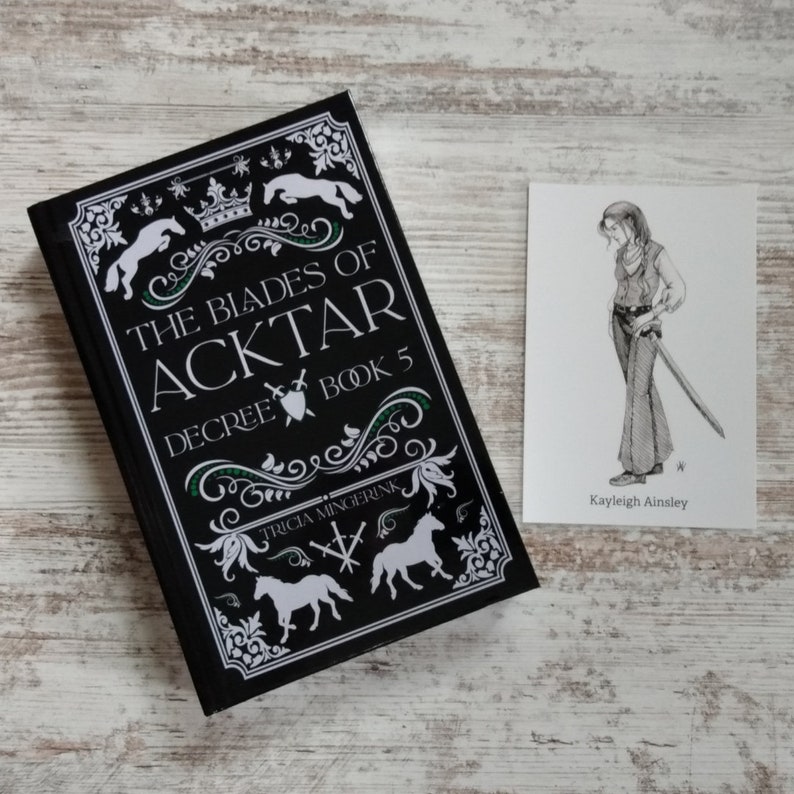 K&ouml;nnte beinhalten: Schwarzes Buch mit wei&szlig;em Text und Illustrationen. Der Titel lautet "The Blades of Aktar, Decree Book 5" von Tricia Mingerink. Eine Schwarzwei&szlig;illustration einer Person mit einem Schwert befindet sich auf einer wei&szlig;en Karte mit dem Text "Kayleigh Ainsley".