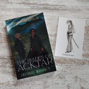 K&ouml;nnte beinhalten: Ein Buch mit dem Titel "The Blades of Acktar, Decree Book 5" von Tricia Mingerink. Der Umschlag zeigt eine gr&uuml;ne Landschaft mit zwei Personen in schwarzer Kleidung. Eine separate Karte mit einer Schwarzwei&szlig;illustration einer Frau, die ein Schwert h&auml;lt, und dem Text "Kayleigh Ainsley".
