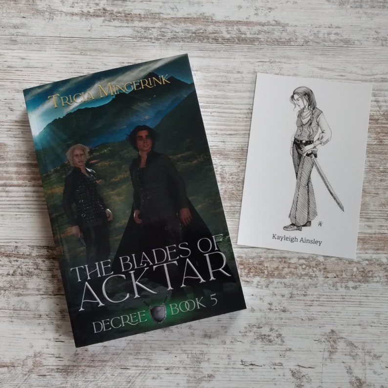 K&ouml;nnte beinhalten: Ein Buch mit dem Titel "The Blades of Acktar, Decree Book 5" von Tricia Mingerink. Der Umschlag zeigt eine gr&uuml;ne Landschaft mit zwei Personen in schwarzer Kleidung. Eine separate Karte mit einer Schwarzwei&szlig;illustration einer Frau, die ein Schwert h&auml;lt, und dem Text "Kayleigh Ainsley".