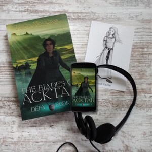 Puede incluir: Un libro de bolsillo titulado "The Blades of Acktar: Defy Book 3" de Tricia Mingerink. La portada muestra a una mujer con una capa negra de pie en un paisaje verde. Un tel&eacute;fono inteligente con la misma portada de libro est&aacute; al lado del libro. Los auriculares est&aacute;n sobre una superficie de madera blanca.