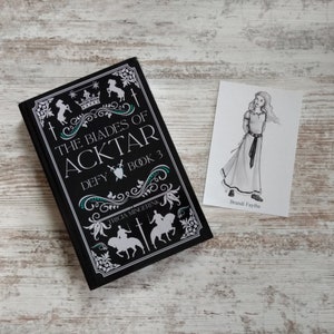 Puede incluir: Un libro negro con un dise&ntilde;o blanco que presenta una corona, espadas y caballos. El t&iacute;tulo es "The Blades of Acktar Defy Book 3" de Tricia Mingerink. Una peque&ntilde;a ilustraci&oacute;n en blanco y negro de una mujer con un vestido largo est&aacute; en una tarjeta blanca con el texto "Brandi Faythe".