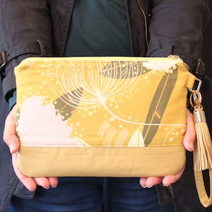 May include: A mustard yellow clutch with a floral print and a brown tassel. The clutch has a zipper closure and a brown leather bottom.