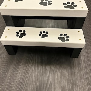 May include: A two-tiered pet step stool. The steps are white with black paw prints and black trim. The steps are made of wood and are designed to help pets reach higher places.