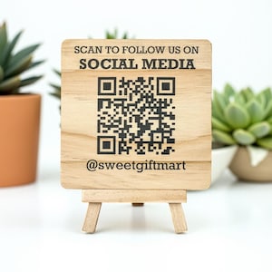 May include: A small wooden sign on a miniature easel. The sign has the text "SCAN TO FOLLOW US ON SOCIAL MEDIA" and a QR code. The sign also includes the username "@sweetgiftmart". The background includes potted succulents.