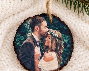 May include: A circular wood slice ornament featuring a printed photograph of a couple embracing. The man wears a dark suit jacket and the woman wears a white off-the-shoulder top with a floral crown. A jute twine loop is attached to the top.