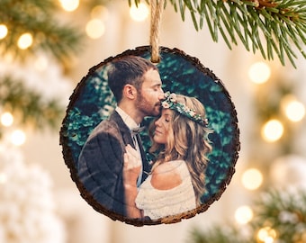 May include: A round wooden slice ornament featuring a printed photograph of a person with short brown hair in a dark suit jacket embracing a person with long wavy blonde hair wearing a white top and a floral crown, hung by a jute twine loop.
