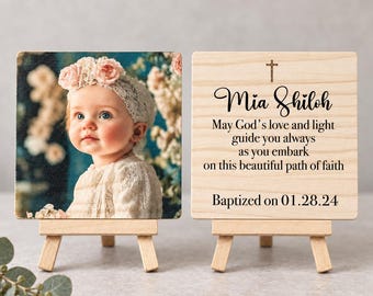 May include: Two square wooden plaques on small easels. One plaque features a photograph of a baby wearing a lace headband with pink flowers. The other plaque displays a cross icon and the text: Mia Shiloh, May God's love and light guide you always as you embark on this beautiful path of faith, Baptized on 01.28.24.
