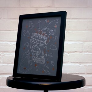 May include: A framed print with a black frame and a gray background. The print features a white line drawing of a metal hand with a symbol on the back of the hand. The hand is surrounded by white line drawings of crystals and stars. The crystals are a light orange color.