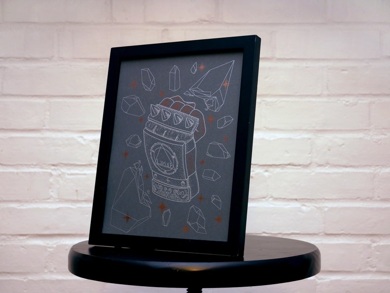 May include: A framed print with a black frame and a gray background. The print features a white line drawing of a metal hand with a symbol on the back of the hand. The hand is surrounded by white line drawings of crystals and stars. The crystals are a light orange color.