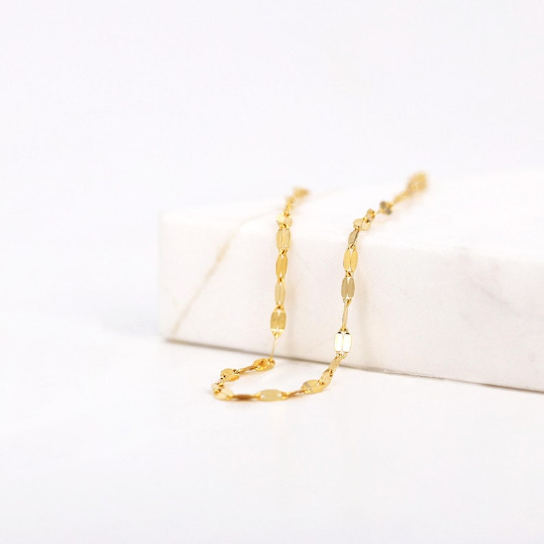 Puede incluir: Un collar de cadena de oro con un delicado dise&ntilde;o de eslabones ovalados. La cadena se muestra sobre una superficie blanca.