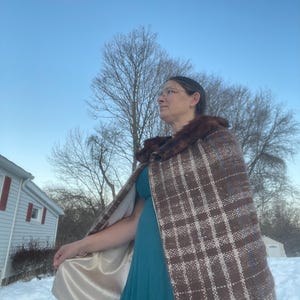 May include: A brown and white plaid cape with a faux fur collar. The cape has a satin lining and is worn over a teal dress. The background shows a snowy landscape and a clear blue sky.