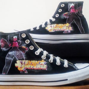 Op de afbeelding: Zwarte hoge sneakers met een kleurrijke afbeelding van een personage uit de animatieserie "Steven Universe". De sneakers hebben witte veters, een witte rubberen neus en het logo van de show. De schoenen worden op een zwarte doos getoond.