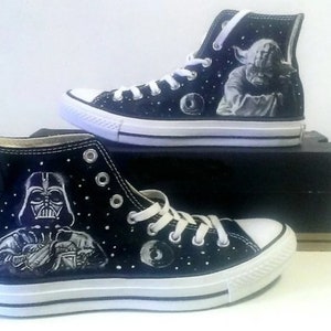 Op de afbeelding: Zwarte hoge sneakers met witte zolen en veters. Eén schoen heeft een Darth Vader-illustratie, de andere Yoda. Beide schoenen hebben een sterrennachtontwerp. Een uniek, handgeschilderd ontwerp voor Star Wars-fans.