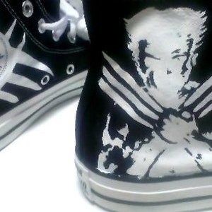 Puede incluir: Zapatillas Converse altas negras con detalles blancos. Una zapatilla presenta el logotipo de Converse All Star con un diseño de estrella. La otra muestra un gráfico blanco de un personaje. Cordones blancos y suela de goma blanca.