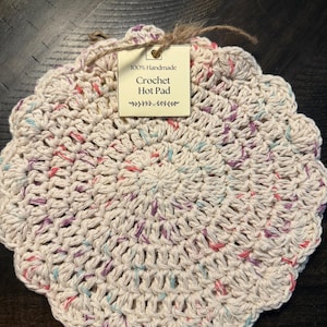 Puede incluir: Un salvamanteles de ganchillo redondo en color blanco roto con borde festoneado y motas multicolores en rosa, morado y azul. Una etiqueta de papel rectangular sujeta con cordel dice 100% Handmade Crochet Hot Pad con un adorno.