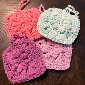 May include: Four square crochet scrubbers featuring a central floral pattern and a small loop for hanging. The set includes one bright pink, one light blue, one purple, and one light lavender scrubber, all crafted with a textured yarn stitch.