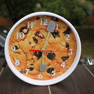 May include: A white round clock with a chocolate chip cookie design. The clock hands are made of wood and have gray metal tips. The numbers on the clock face are black and the clock face has a red second hand.
