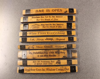 May include: A collection of wooden barrel stave signs with various engraved phrases. The signs are rectangular, with metal bands and dark text. Phrases include "BAR IS OPEN" and "Bourbon May Not Be the Answer".