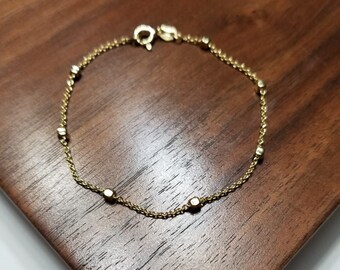 May include: A delicate gold chain bracelet with small round beads. The bracelet is on a wooden surface.