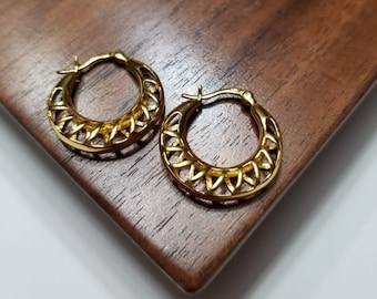 May include: A pair of gold hoop earrings with a geometric design. The earrings are on a wooden surface.